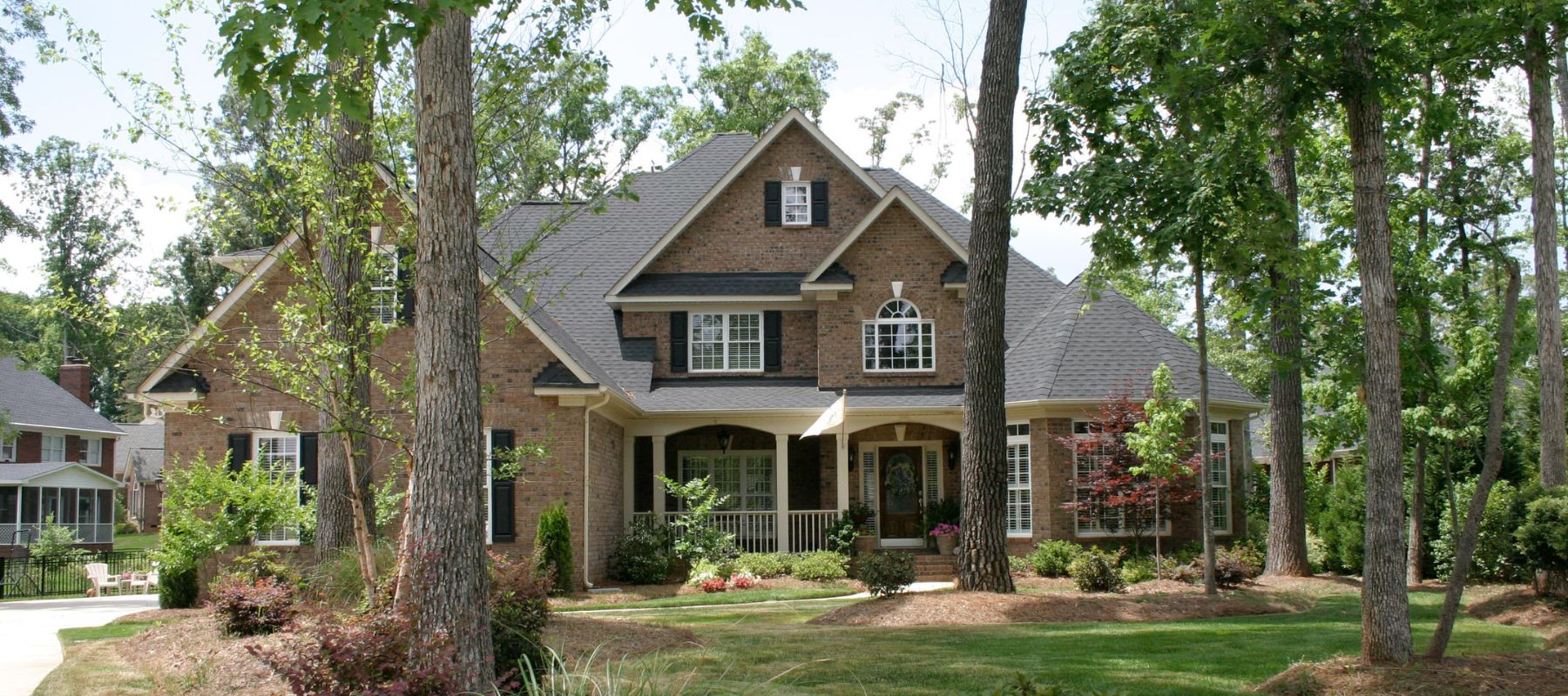 Two-story brick house with black shutters surrounded by mature trees and landscaping