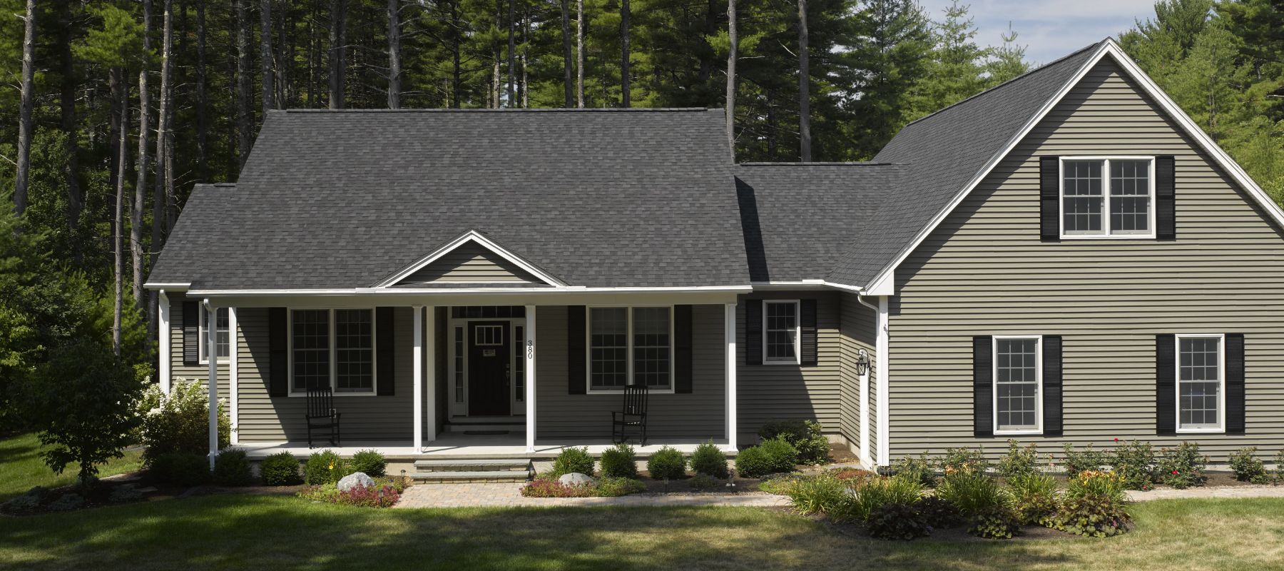 Single-story home with light olive siding, black shutters, and a front porch nestled among tall trees