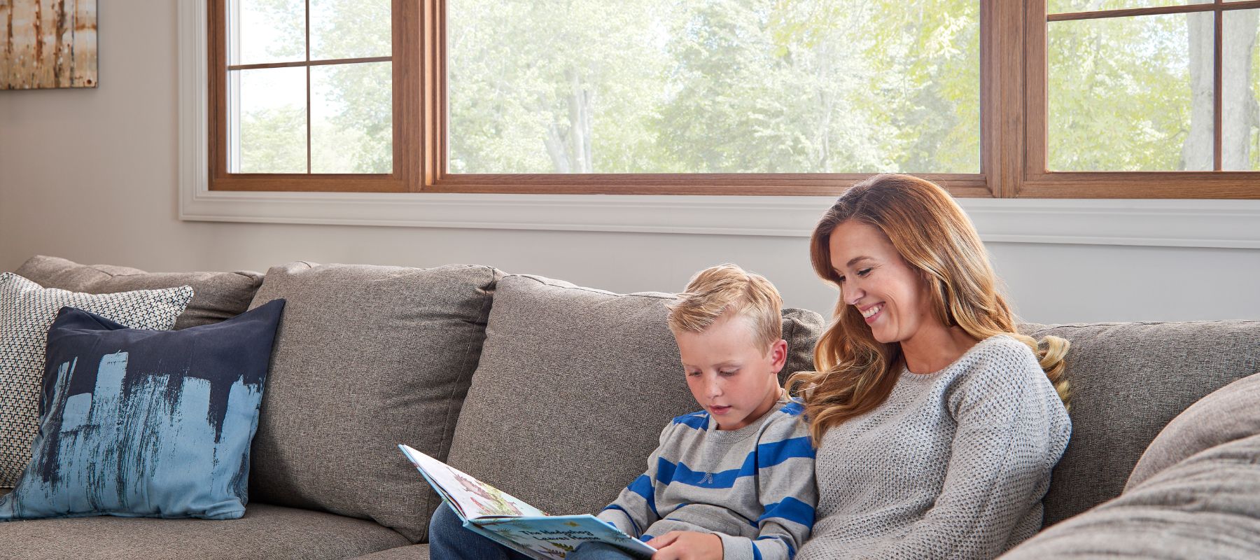 Mother and young son reading a book together on a grey sofa in a bright living room with wood-trimmed windows