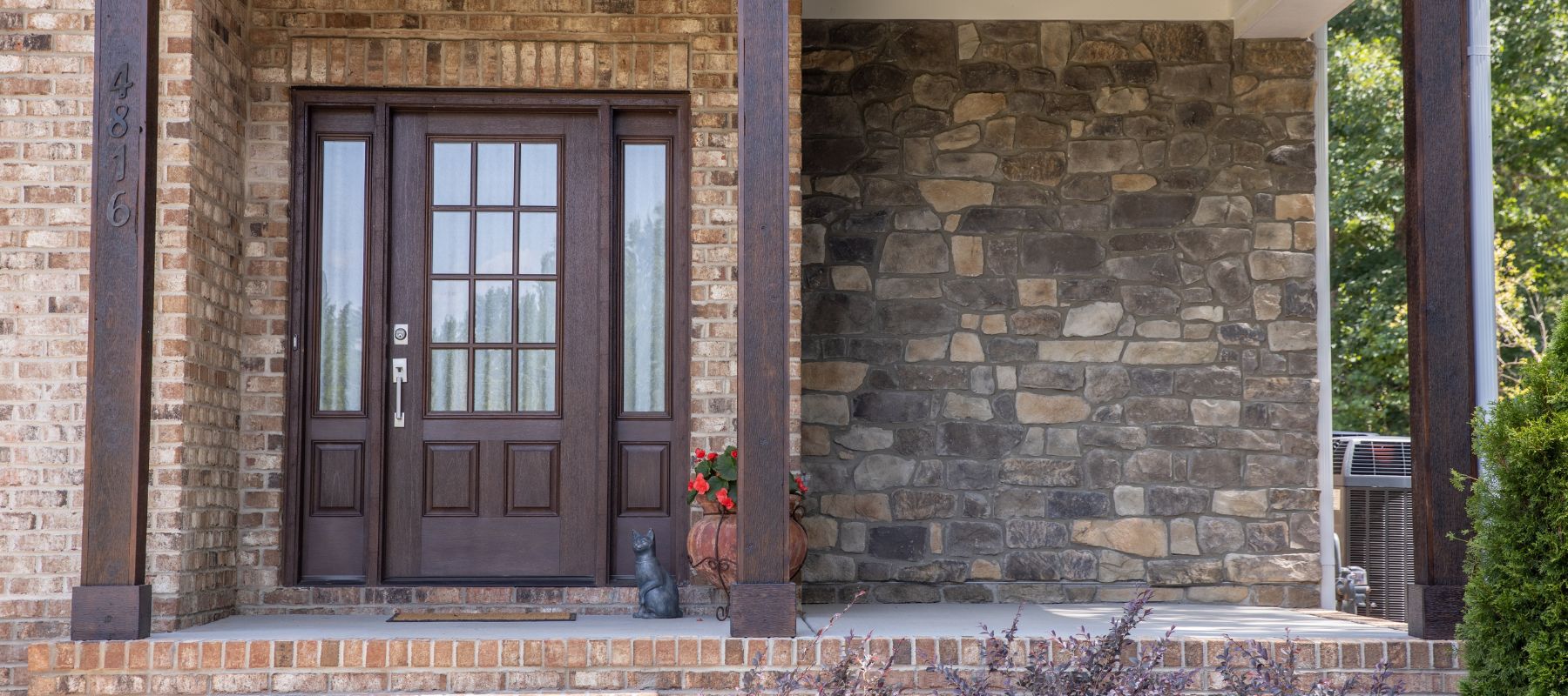 Front porch of a brick house with a dark wood door and stone accent wall, accented by a potted plant and a cat statue