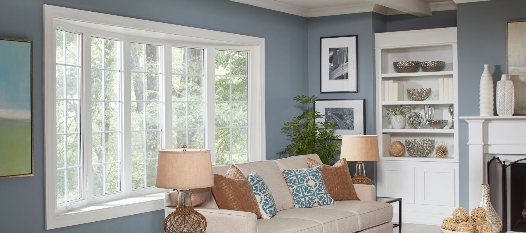 A stylish living room features a large bay window with white grilles looking out onto lush greenery, complemented by a light-colored sofa with throw pillows, lamps, and a built-in bookshelf